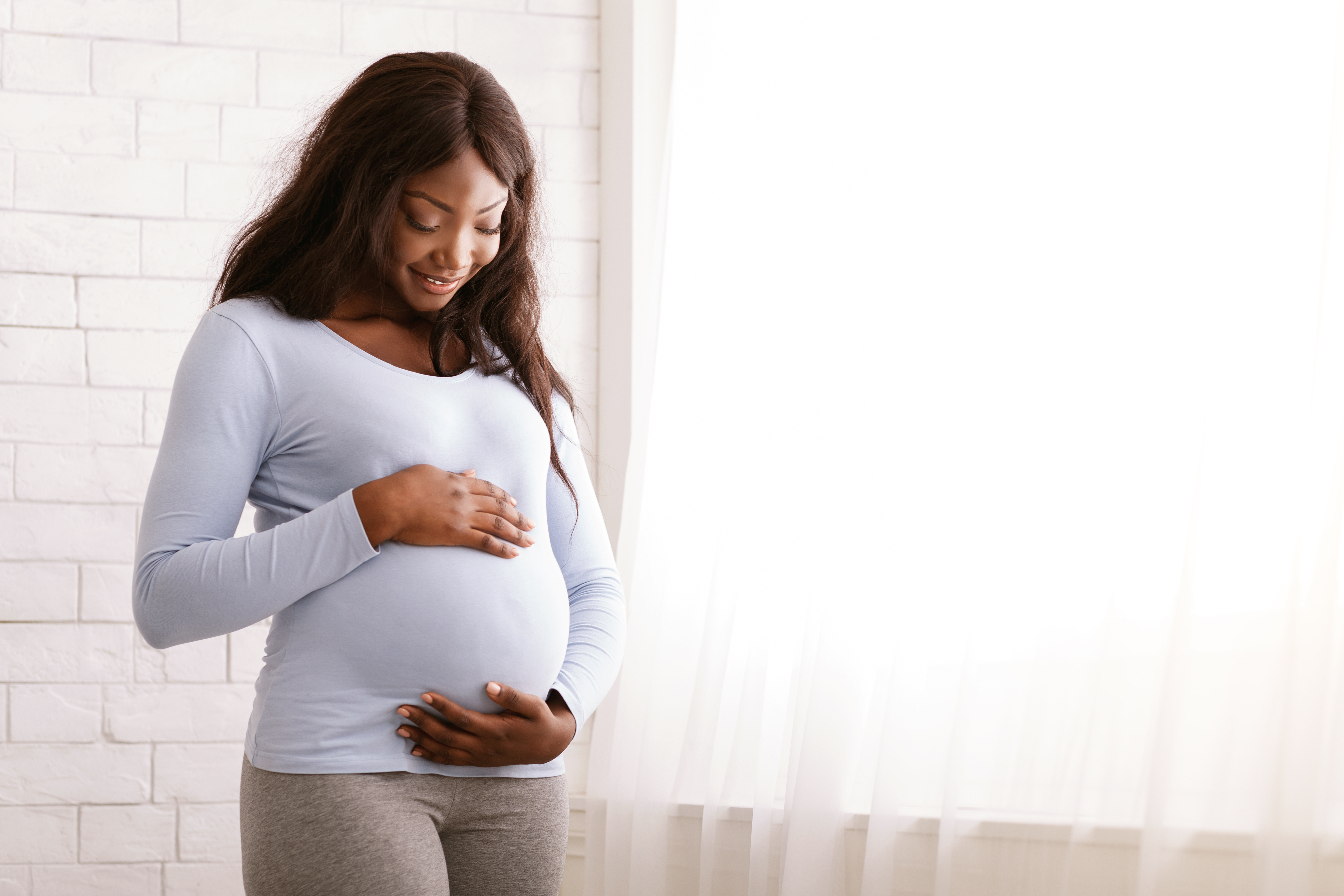 Peaceful pregnant black woman hugging her tummy next to window at home, free space