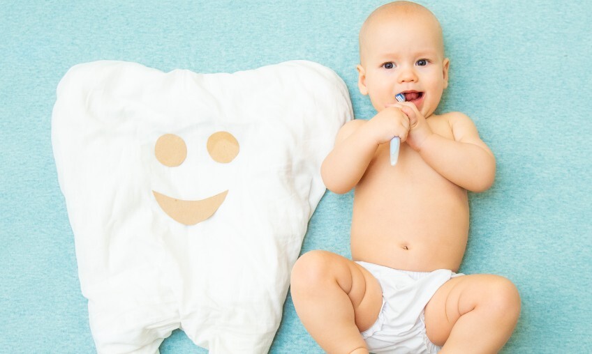 a baby laying on a blue blanket with a white toothbrush it its mouth beside a white pillow shaped like a tooth